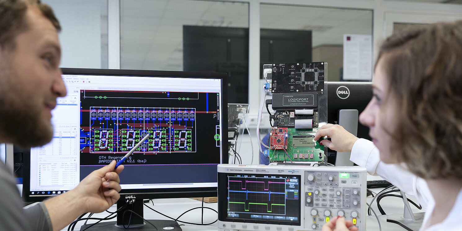 Zwei Studierende besprechen Messergebnisse an einem elektronischen Versuchsaufbau, im Hintergrund eine elektronische Schaltung, ein Computerbildschirm und ein Oszilloskop. 